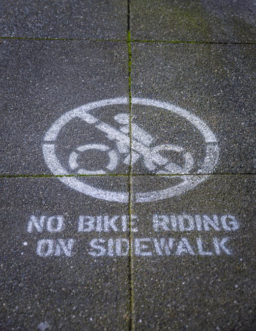 A concrete sidewalk surface featuring a circular white stencil marking with a printed image of a bicycle crossed out by a diagonal line, accompanied by the text 'NO BIKES RIDING ON SIDEWALK' beneath t